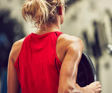 A woman with blonde hair in a bun wears a red tank top, holding a weight plate in a gym setting.