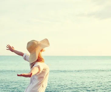 Woman in a hat with arms outstretched, enjoying the ocean view on a sunny day.