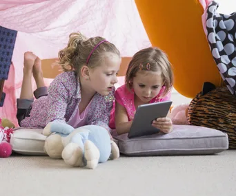 Two young girls lying on cushions in a tent, focused on a tablet.