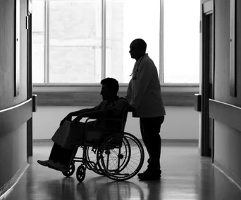 "Silhouette of a healthcare worker pushing a patient in a wheelchair down a brightly lit hospital corridor."