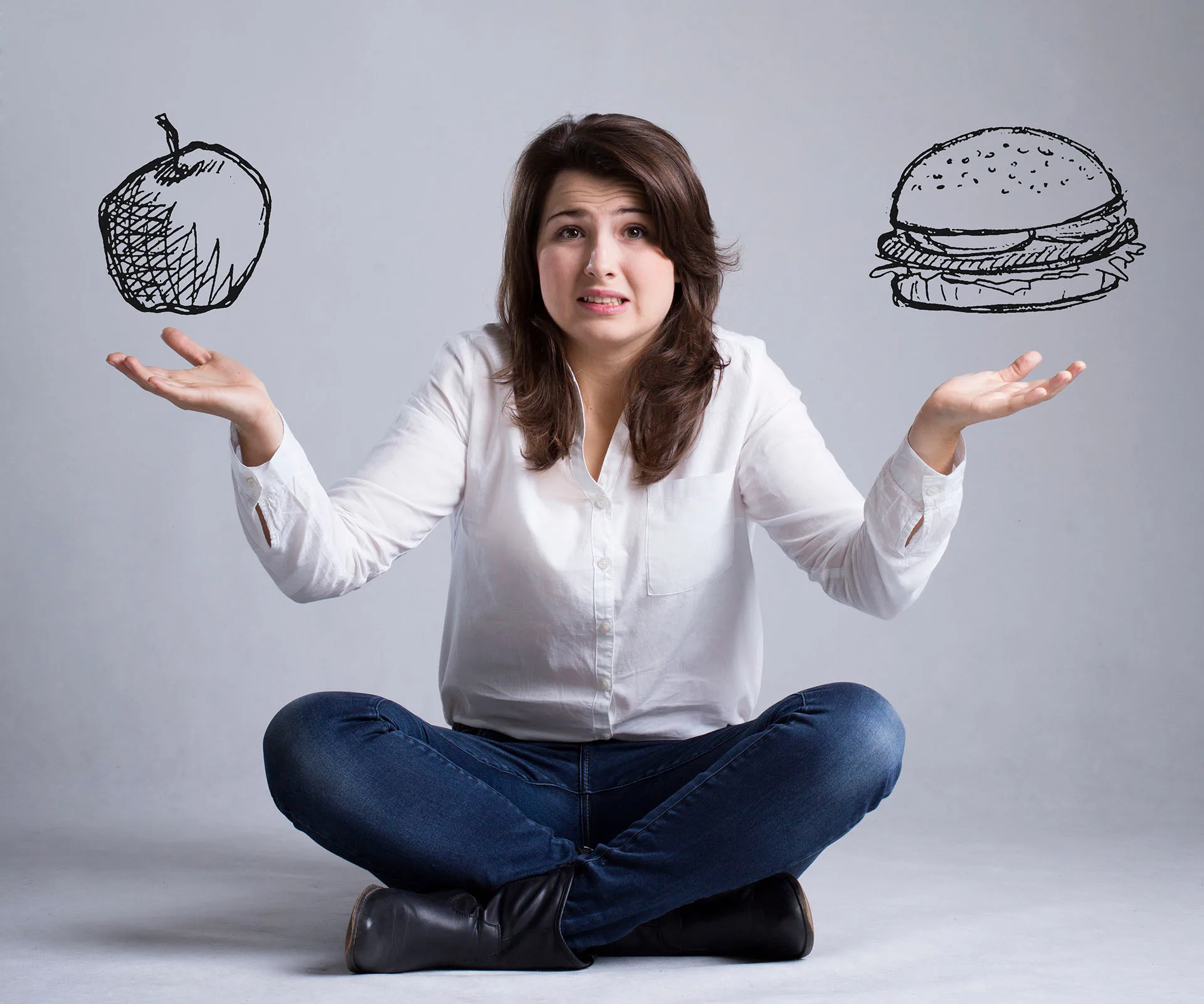 Woman sitting cross-legged, puzzled between a drawn apple and burger on each side, wearing a white shirt and jeans.