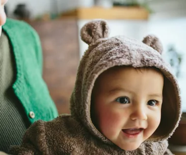 Baby in a cozy, brown bear hoodie smiles widely, with an adult wearing a green sweater next to them.