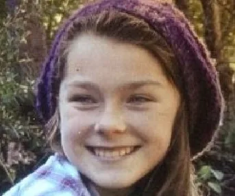 Young person smiling outdoors wearing a purple knit hat, surrounded by greenery.