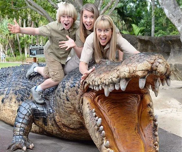Three people cheerfully pose on a large crocodile sculpture in a lush, outdoor setting.