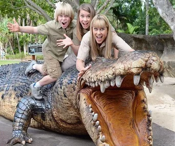 Three people cheerfully pose on a large crocodile sculpture in a lush, outdoor setting.