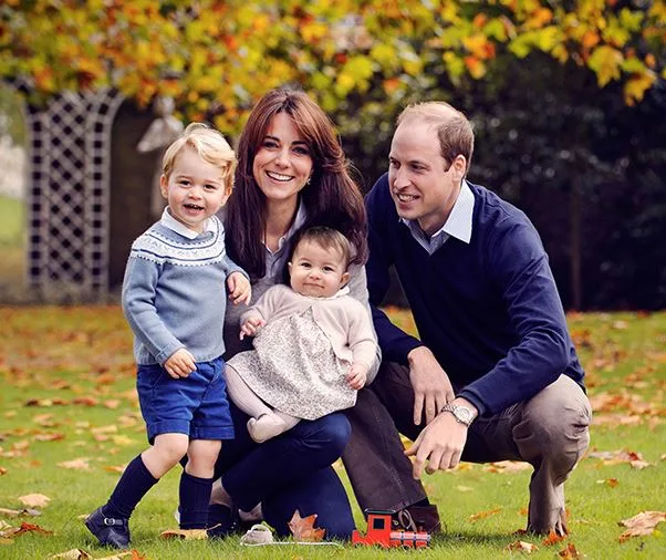A family of four, including two adults and two young children, pose together on a lawn with autumn leaves around them.