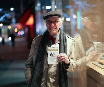 Elderly man smiling in a coat and hat, holding Shakespeare's "The Sonnets" book, standing on a city street at night.