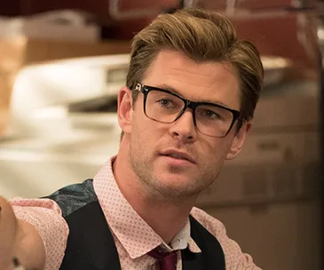 A man with glasses, styled hair, and formal attire appears to be in an office setting.