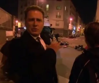 A man in a suit on a dimly lit street gestures towards a distant scene with emergency vehicles and scattered debris.