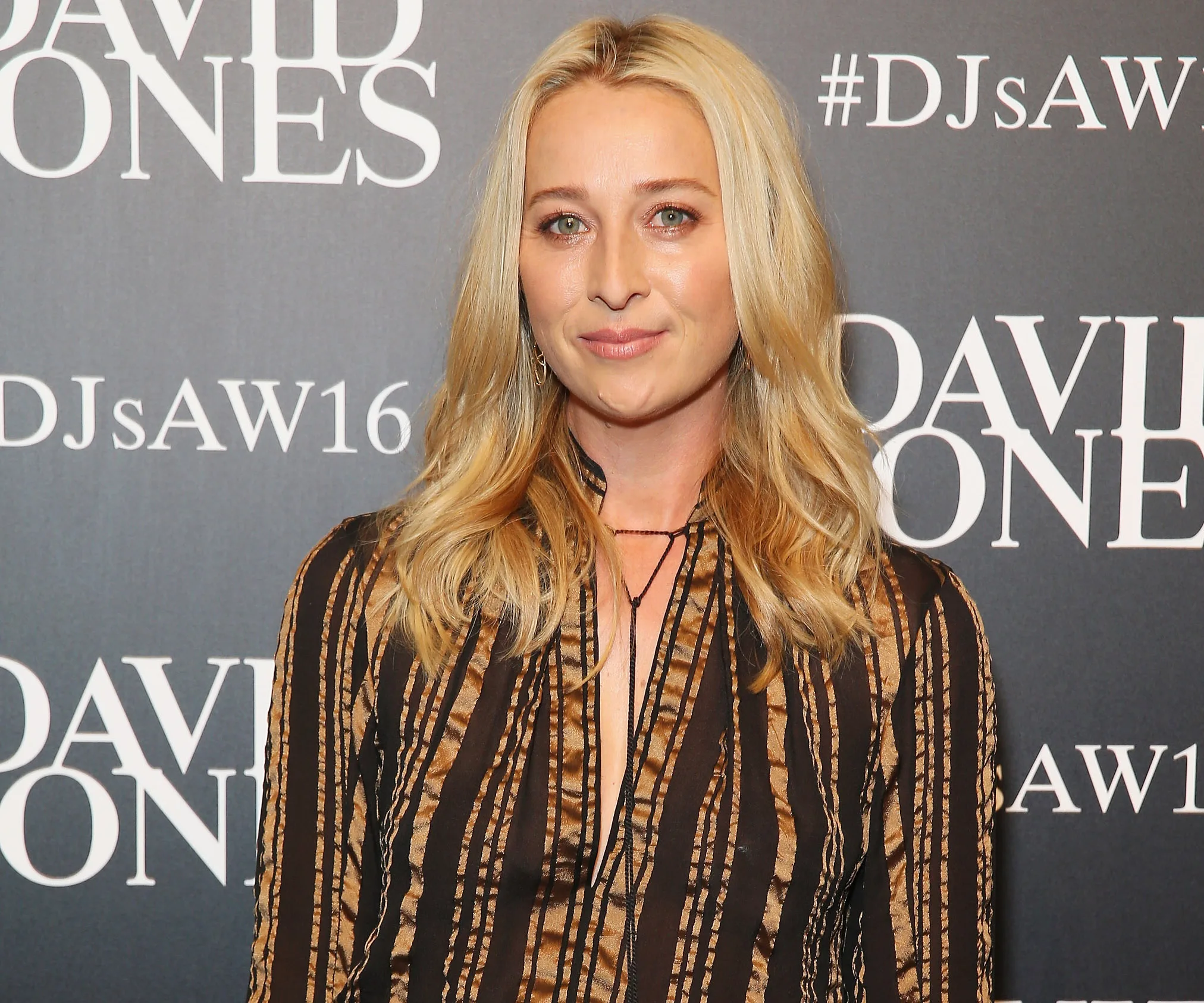 A person with long blonde hair wearing a striped outfit, standing in front of a David Jones backdrop at an event.