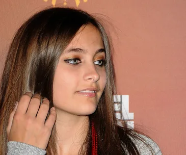Young woman with long hair and makeup, looking off-camera, against a warm-toned background.