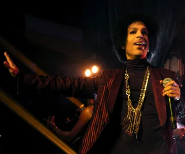 Musician in a vibrant outfit performing on stage, holding a microphone, with dramatic lighting in the background.