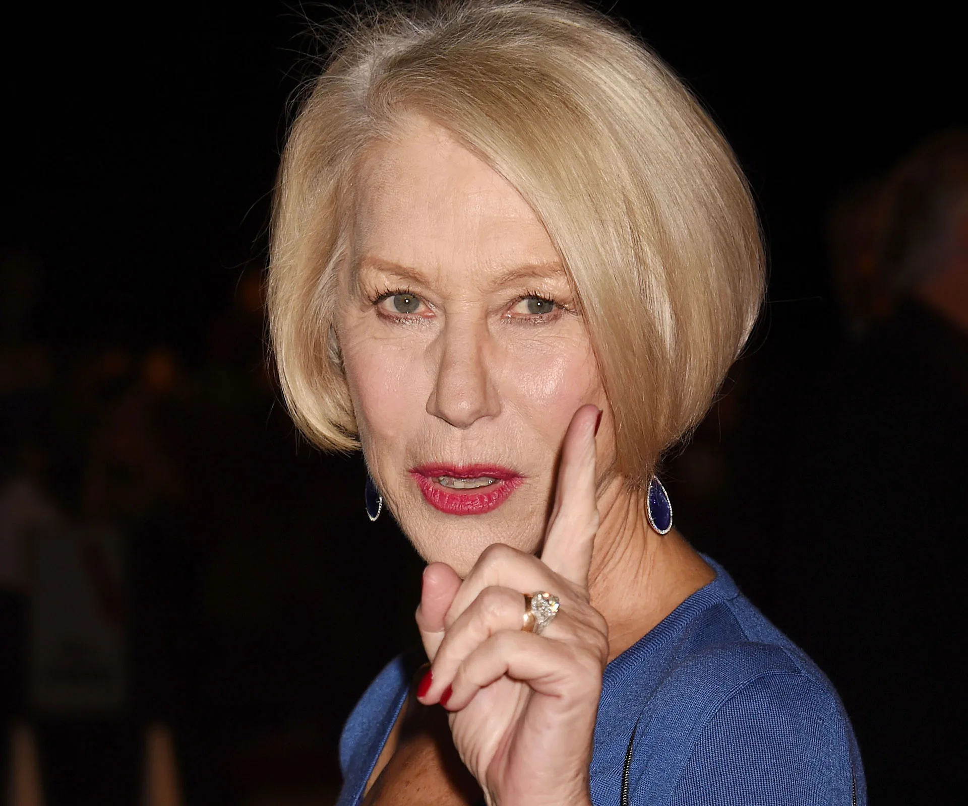 An older woman with short blonde hair and blue attire gestures with her finger, conveying emphasis.