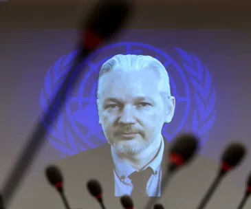 Julian Assange appears on a screen with a United Nations emblem in the background, framed by multiple microphones.