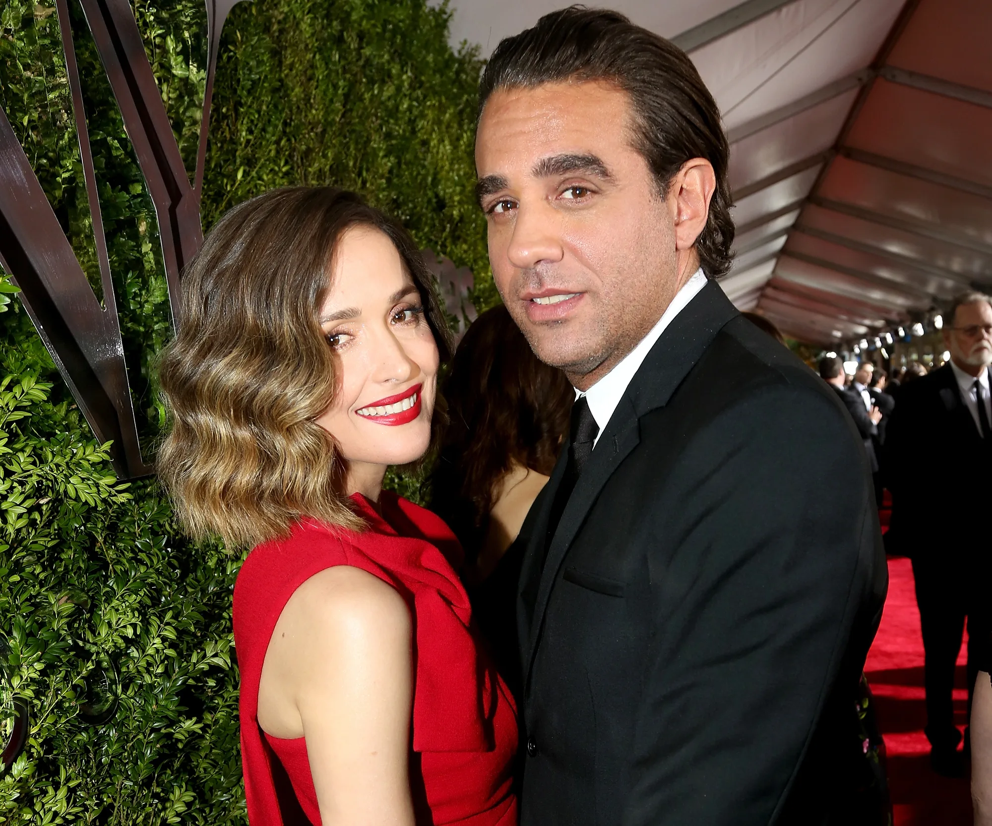 Two people pose and smile on a red carpet, surrounded by other guests and greenery.
