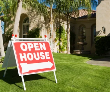 "Red open house sign on lush lawn with palm trees and elegant home in background."