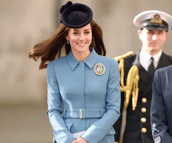 A woman in a blue coat with a diamond brooch and a navy hat, smiling outdoors.