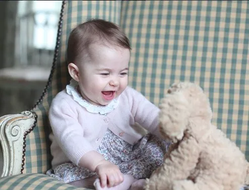 A baby wearing a pink sweater sits on a chair, smiling at a stuffed teddy bear.