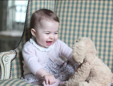 A baby wearing a pink sweater sits on a chair, smiling at a stuffed teddy bear.