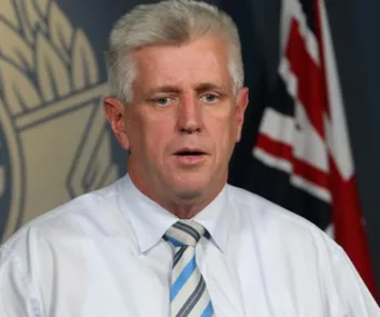 A man in a white shirt and striped tie stands in front of a flag and emblem backdrop, with a serious expression.