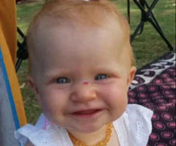 A baby smiling outdoors, sitting on grass near a patterned blanket, wearing a white outfit and a yellow necklace.