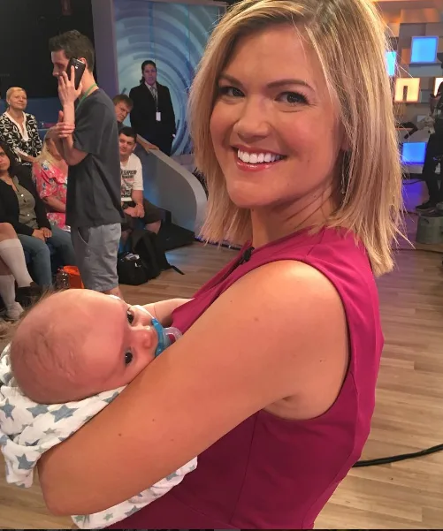 A woman in a pink dress smiling while holding a baby in a studio setting with people in the background.