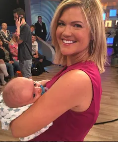 A woman in a pink dress smiling while holding a baby in a studio setting with people in the background.