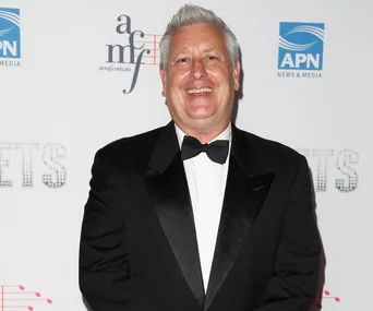 A smiling man in a tuxedo at a media event with a white backdrop featuring logos.