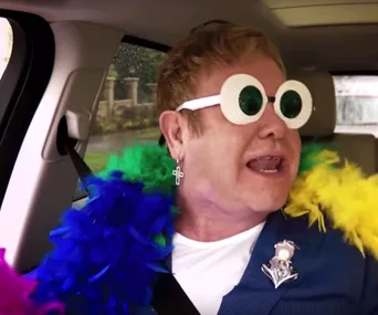 Elton John in colorful boa and quirky glasses singing in a car.