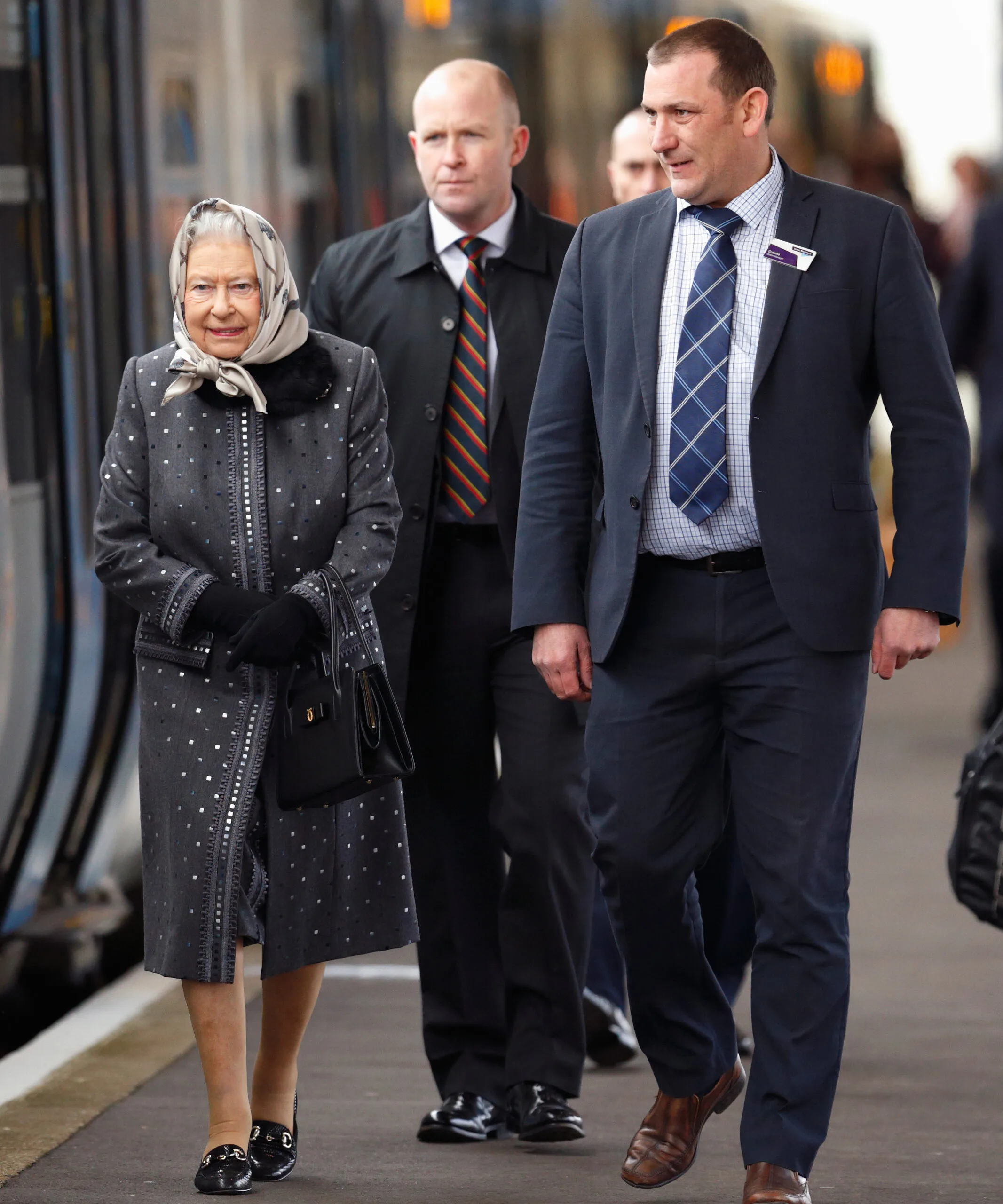 Queen walking on a train platform with two men, wearing a scarf and coat.