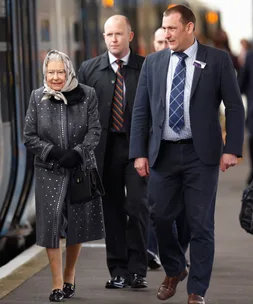 Queen walking on a train platform with two men, wearing a scarf and coat.