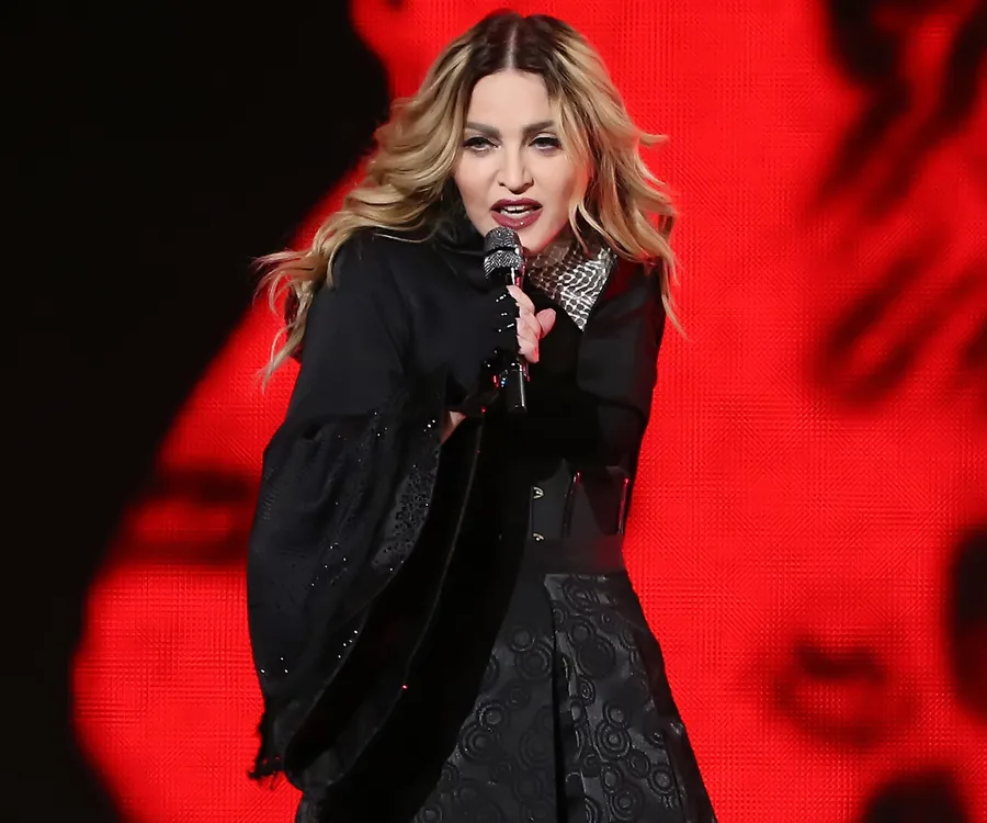 Singer with long hair performs energetically on stage, holding a microphone against a vibrant red background.