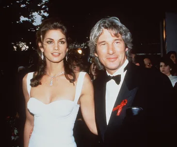 A couple poses elegantly in evening attire, the woman in a white gown and the man in a black tuxedo with a red ribbon.