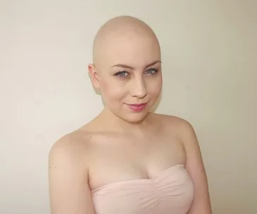 A young woman with a shaved head and a soft smile wearing a strapless top, against a plain background.