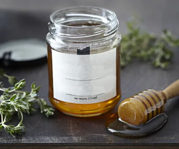 Open jar of honey beside a honey dipper, with fresh herbs in the background on a wooden surface.
