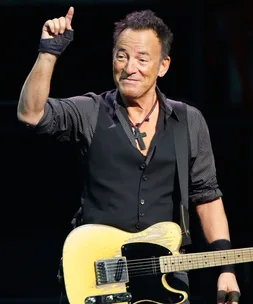 Musician in a dark vest and shirt holding a yellow electric guitar, giving a thumbs-up on stage.