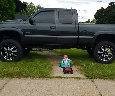 A baby in a small toy car under a large, lifted black truck on a driveway.
