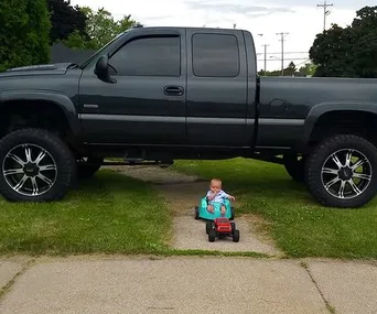 A baby in a small toy car under a large, lifted black truck on a driveway.
