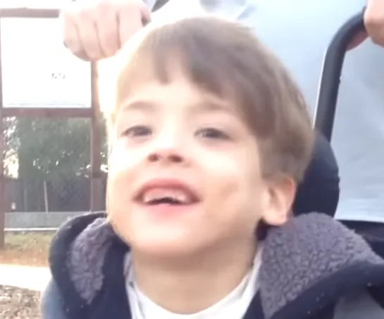 Dad pushes smiling son in wheelchair at skate park on a sunny day.