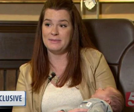 A woman with long hair holds a sleeping baby in her arms during an interview, labeled "EXCLUSIVE" on screen.