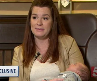 A woman with long hair holds a sleeping baby in her arms during an interview, labeled "EXCLUSIVE" on screen.