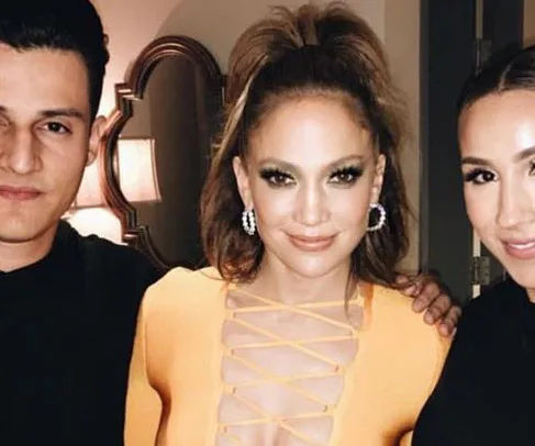 Jennifer Lopez in a yellow lace-up dress, posing with two people, smiling.