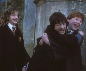 Three young characters from Harry Potter, in school robes, laughing and embracing outside near a stone wall.