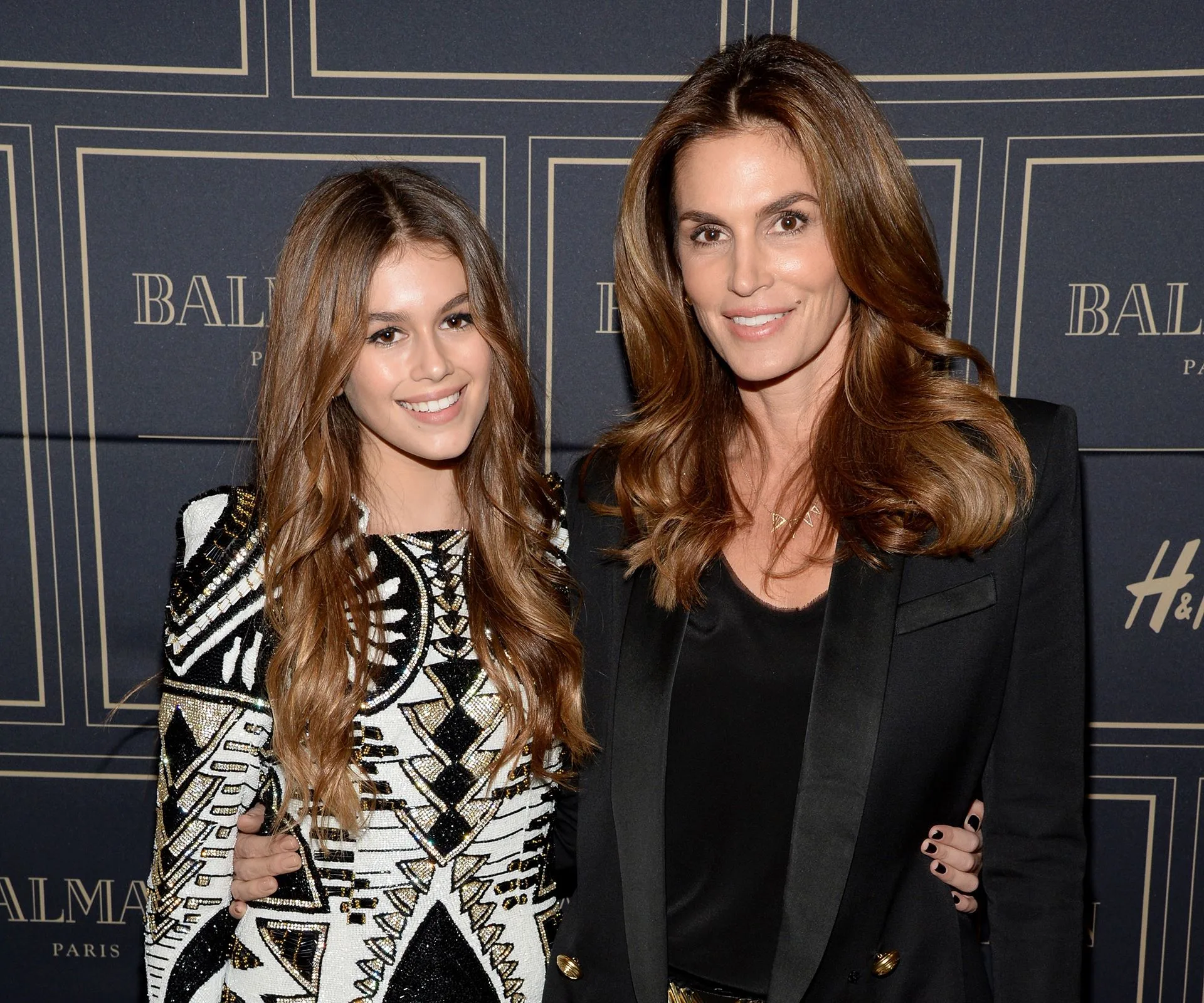 Mother and daughter in stylish outfits at a fashion event backdrop, smiling and posing together.
