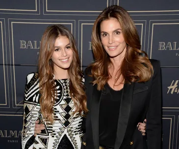 Mother and daughter in stylish outfits at a fashion event backdrop, smiling and posing together.