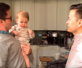 A baby looks confused between two similar-looking adults in a kitchen, making it unclear who the dad is.