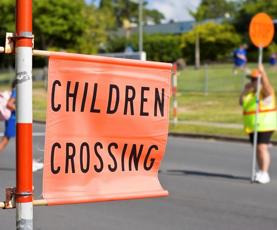 "Orange 'Children Crossing' sign near road with blurred school lollipop lady in the background holding a stop sign."
