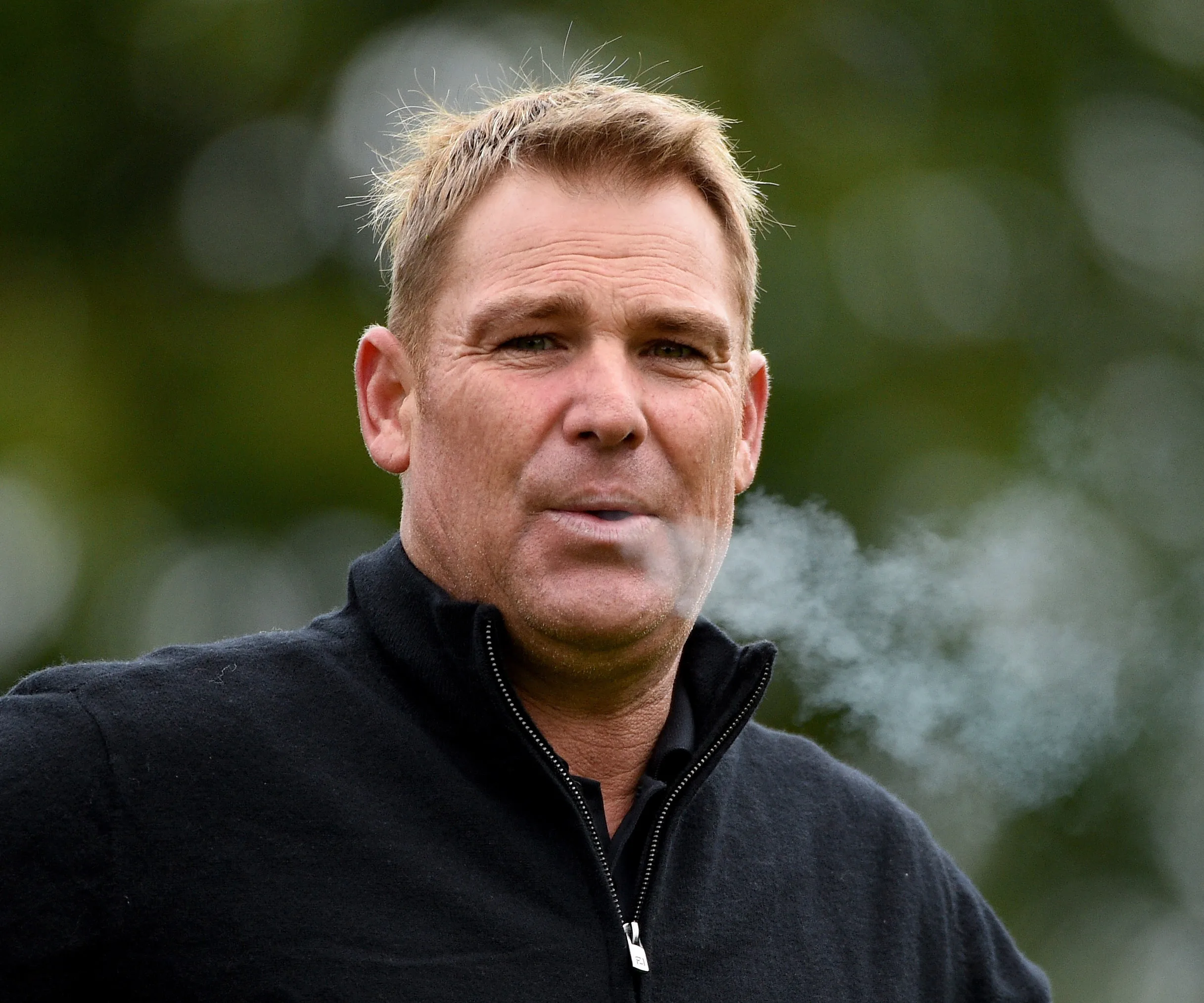 Man in a black jacket outdoors, exhaling smoke.