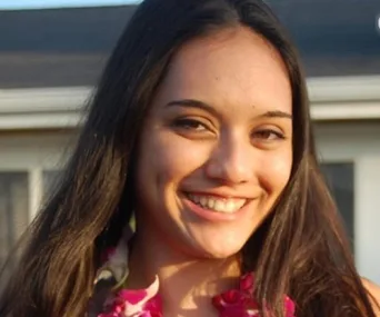 A young woman with long dark hair smiles warmly, wearing a floral-patterned top, standing in front of a house.