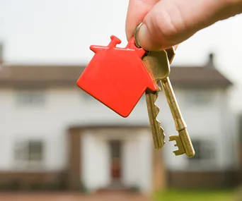 Hand holding keys with a red house-shaped keychain, blurry house in background.